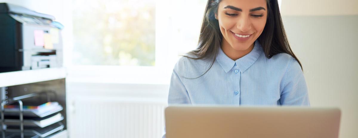 Young woman, remote employee working at laptop in a home office, with a desktop or multifunction printer in the background